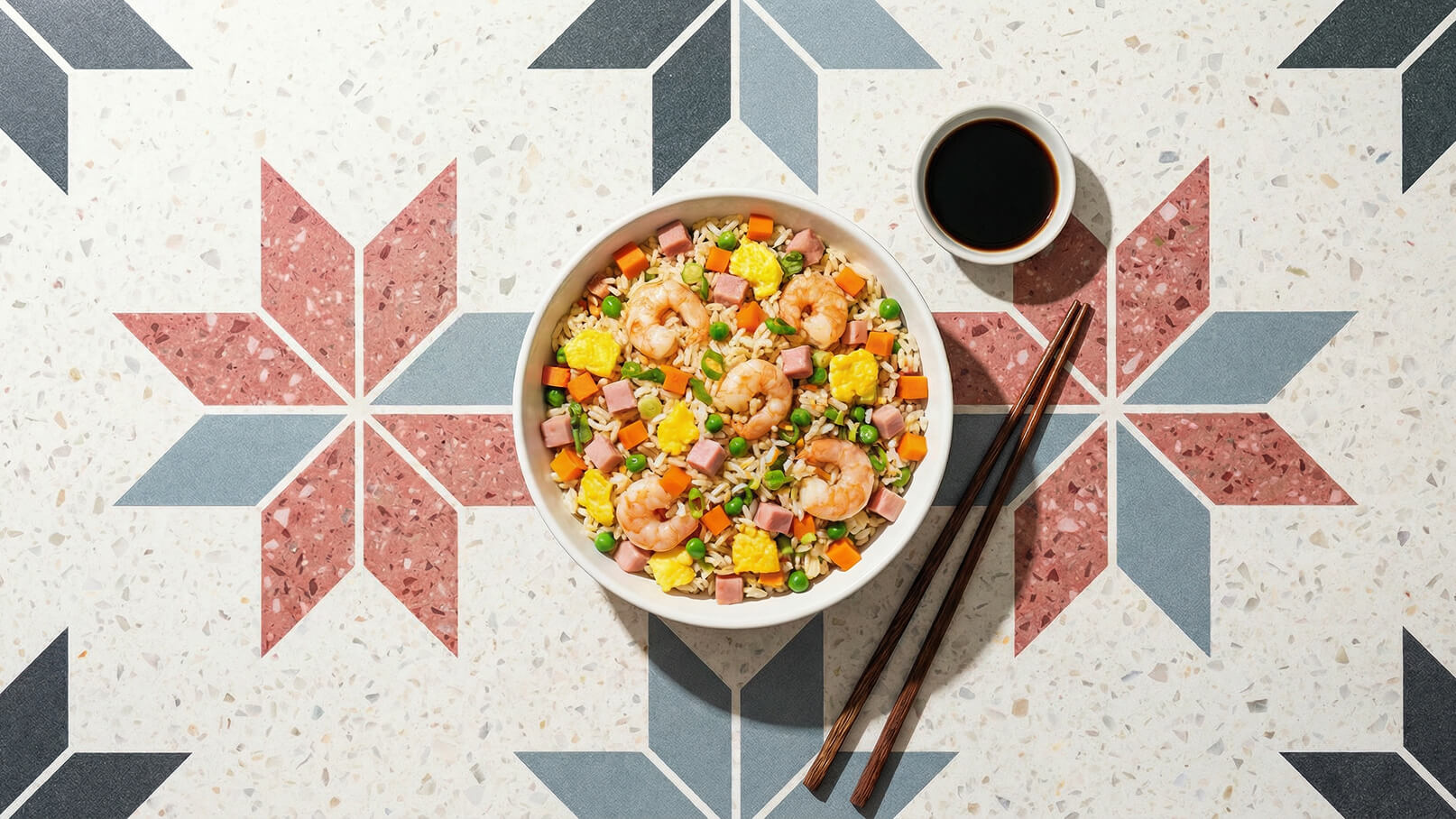 Eine Schüssel mit gebratenem Reis, garniert mit Garnelen, buntem Gemüse und Eiern. Neben der Schüssel liegen Essstäbchen und eine kleine Schüssel mit Sojasauce. Der Hintergrund zeigt ein dekoratives Muster in verschiedenen Farben.