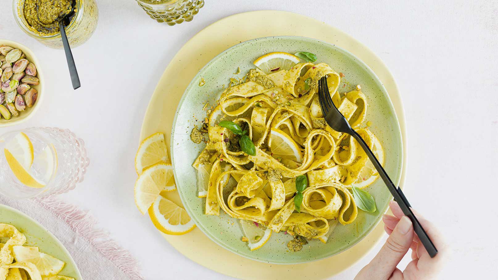 Ein Teller mit frischen, gelben Tagliatelle, garniert mit grünem Pesto und Basilikumblättern. Neben dem Teller sind Zitronenscheiben und eine Gabel sichtbar. Im Hintergrund ist ein Glas mit Wasser und Zitrone zu sehen.