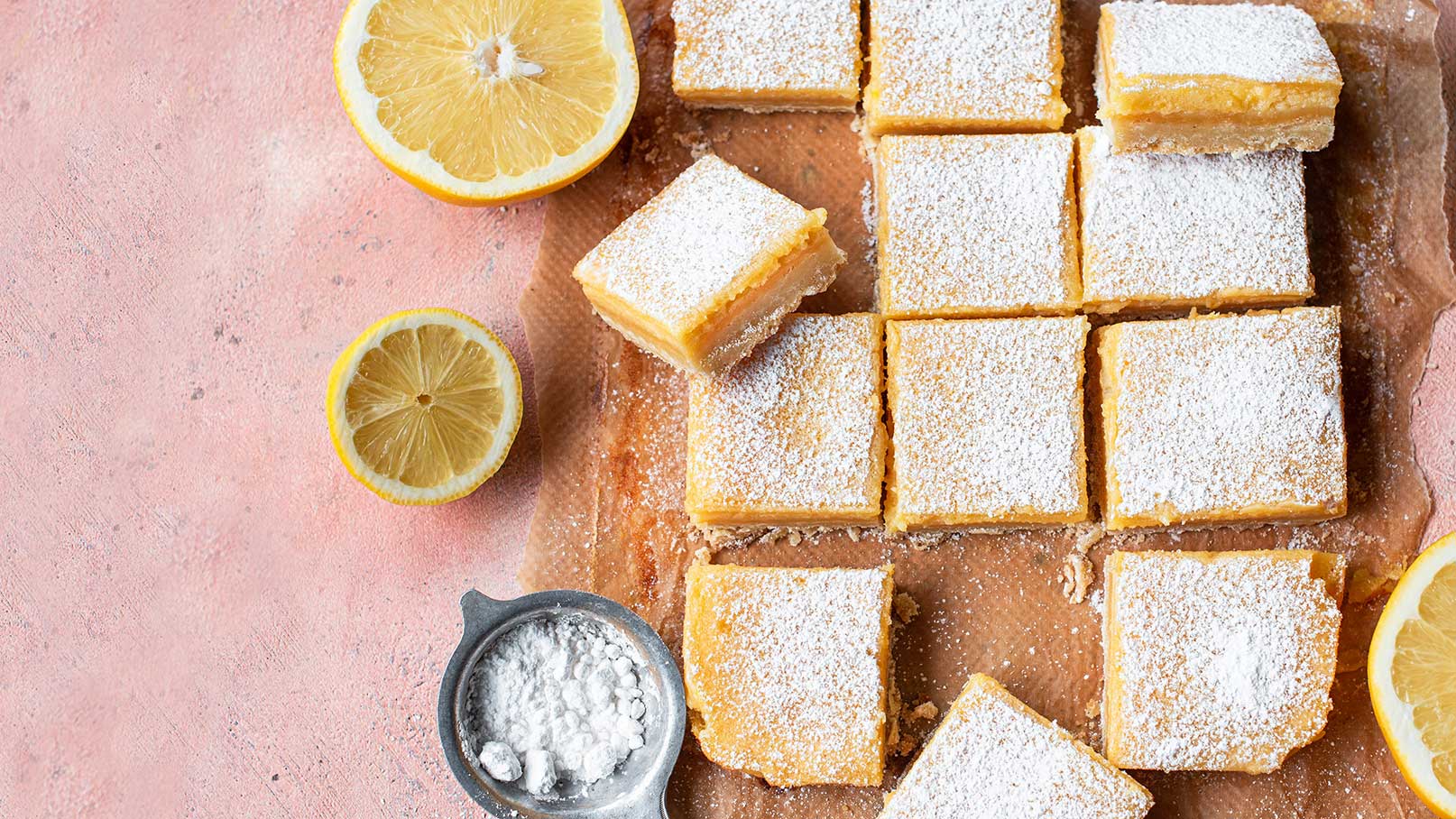 Ein Tablett mit frisch gebackenem Zitronenkuchen, in gleichmäßige Stücke geschnitten und mit Puderzucker bestäubt. Neben dem Kuchen liegen zwei halbierte Zitronen. Ein kleines Schälchen mit Puderzucker befindet sich ebenfalls auf dem Tablett. Der Hintergrund ist blassrosa.