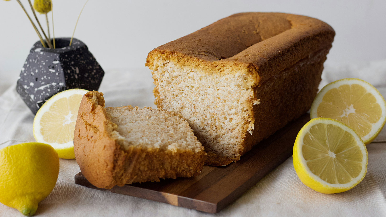 Ein frisch gebackener Zitronenkuchen liegt auf einem Holzbrett. Ein Stück des Kuchens ist bereits geschnitten. Neben dem Kuchen liegen zwei halbierte Zitronen. Der Hintergrund ist neutral gehalten, um den Fokus auf den Kuchen und die Zitronen zu lenken.