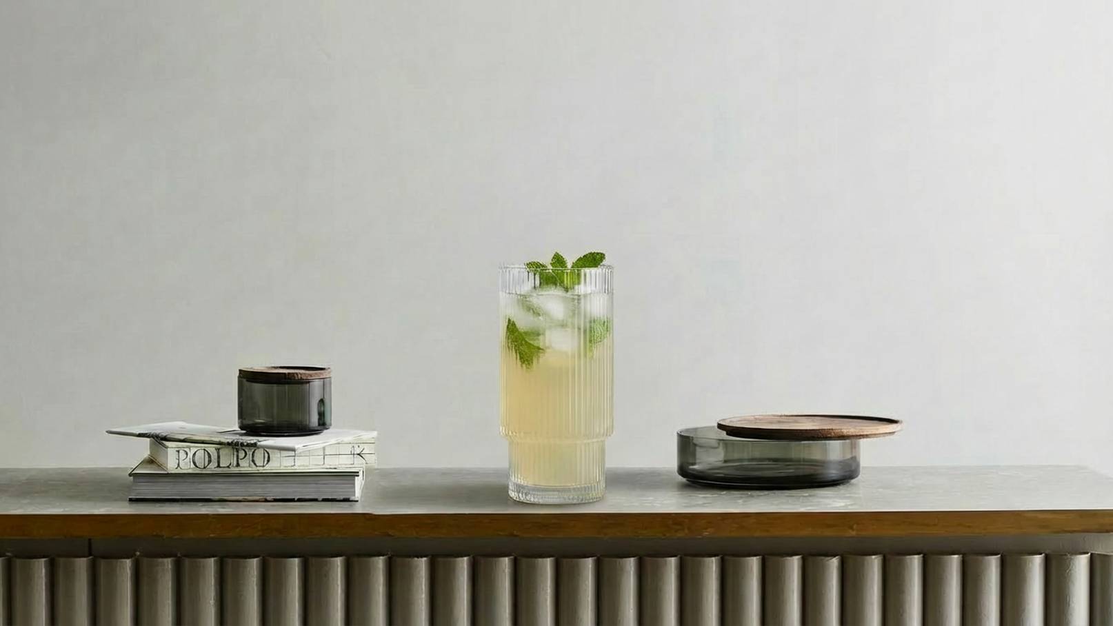 Ein dekorativer Tisch mit einem hohen Glas, das ein kühles Getränk mit Minzblättern enthält. Daneben steht eine kleine schwarze Tasse und ein Stapel Bücher. Rechts liegt ein flacher Teller. Der Hintergrund ist hell und neutral.