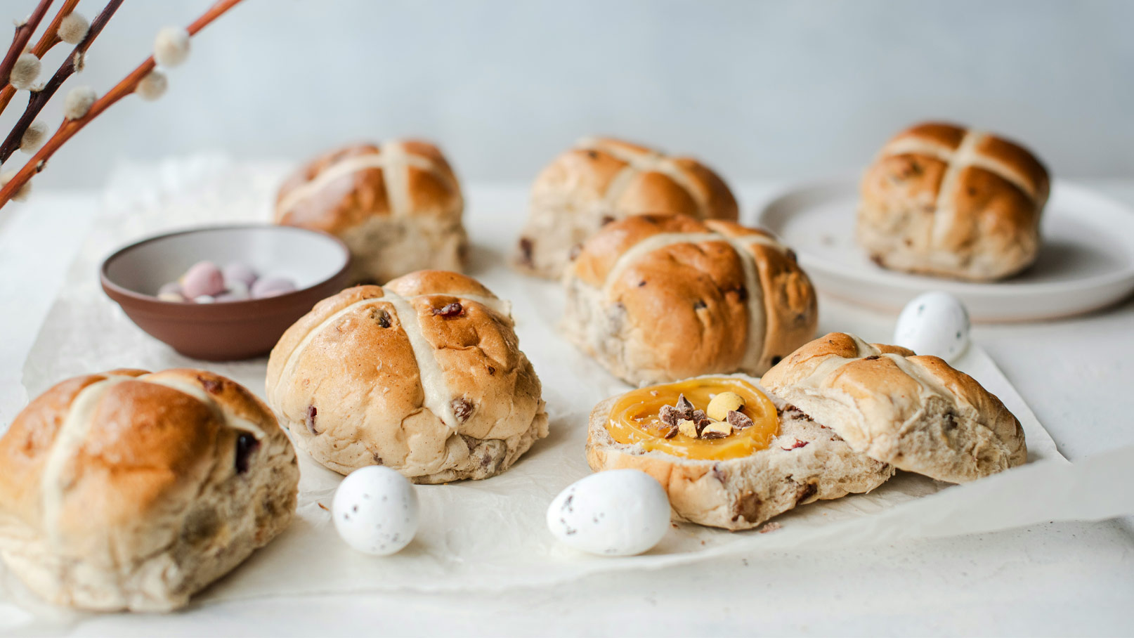 Eine Auswahl von frischen Hot Cross Buns auf einem Tisch. Einige Brötchen sind ganz, während eines aufgeschnitten ist, mit einer gelben Füllung und Schokoladensplittern. Im Hintergrund liegt eine Schüssel mit bunten Schokoladen-Eiern.
