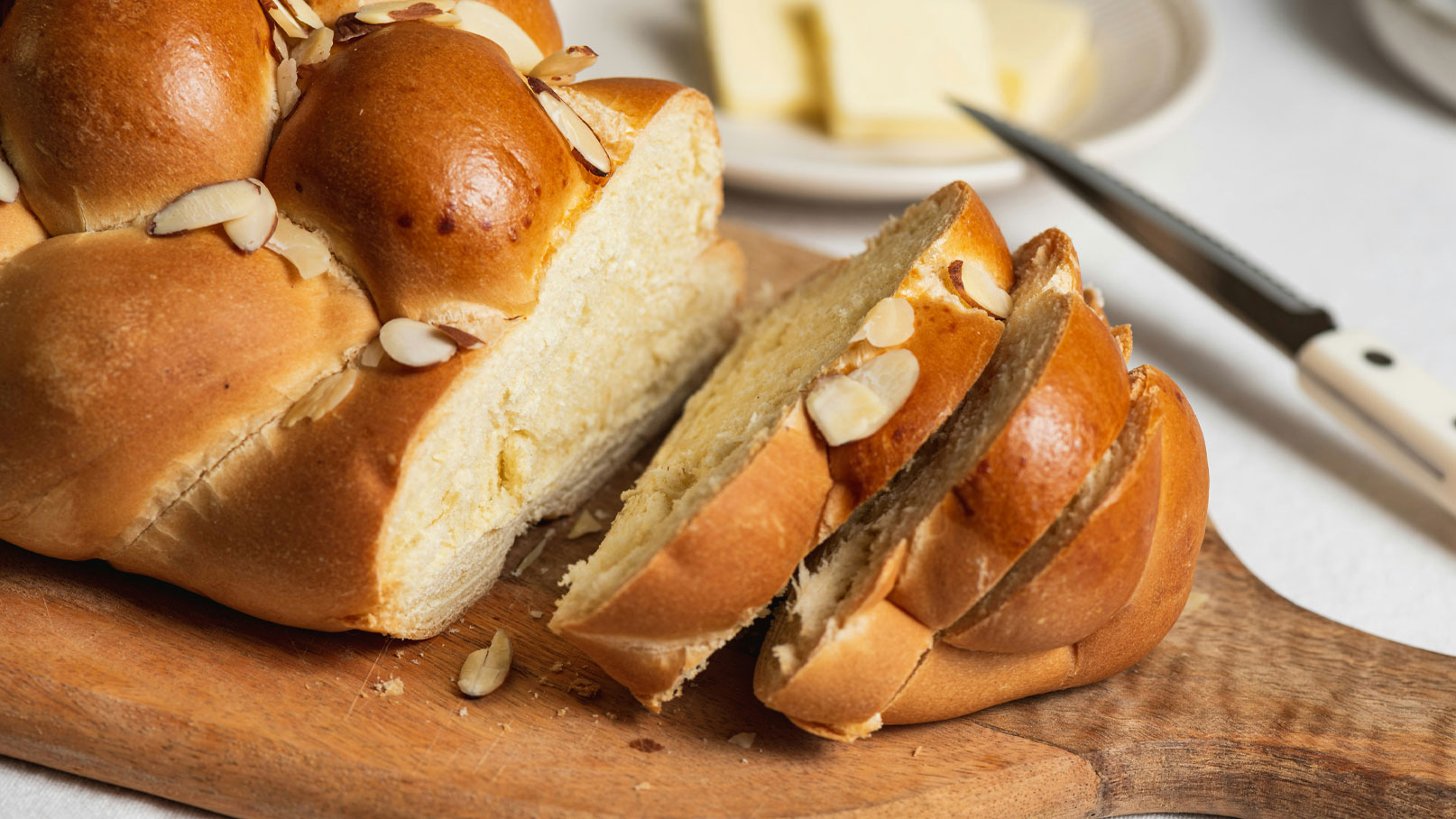 Ein frisch gebackenes, geflochtenes Brot mit einer goldbraunen Kruste und gehobelten Mandeln obendrauf liegt auf einem Holzbrett. Ein Teil des Brotes ist angeschnitten und zeigt die weiche, helle Krume. Im Hintergrund sind Butterstücke auf einem Teller zu sehen.