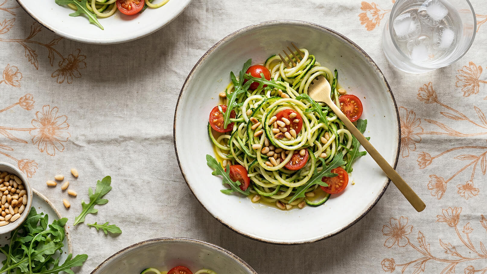 Ein Teller mit Zoodles (Zucchini-Nudeln) ist mit Kirschtomaten, frischem Rucola und gerösteten Pinienkernen garniert. Eine goldene Gabel liegt auf dem Teller, während ein Glas Wasser nebenan steht. Der Tisch ist mit einer blumigen Tischdecke gedeckt.