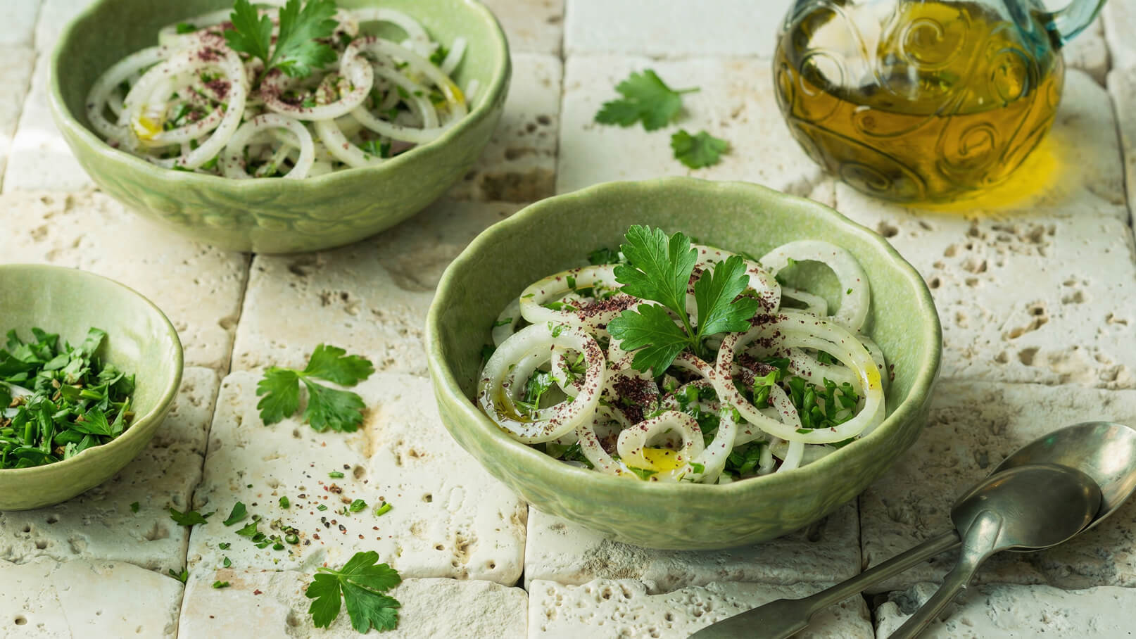 Eine grüne Schüssel mit einem frischen Salat aus Zwiebelringen, Kräutern und Gewürzen. Ein kleinerer, ähnlicher Teller enthält ebenfalls gehackte Kräuter. Im Hintergrund steht eine dekorative Ölkaraffe. Der Tisch ist mit hellen, strukturierten Fliesen bedeckt.