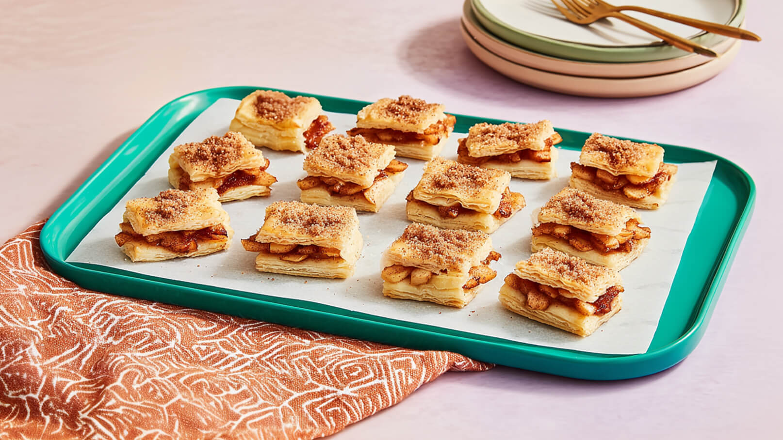 Auf einem grünen Tablett liegen mehrere schichtweise gebaute Gebäckstücke, gefüllt mit Äpfeln und bestreut mit Zimt-Zucker. Die Gebäckstücke sind gleichmäßig angeordnet und auf Backpapier platziert. Im Hintergrund ist ein stilvoller Tisch mit einer Anleitung zu sehen.