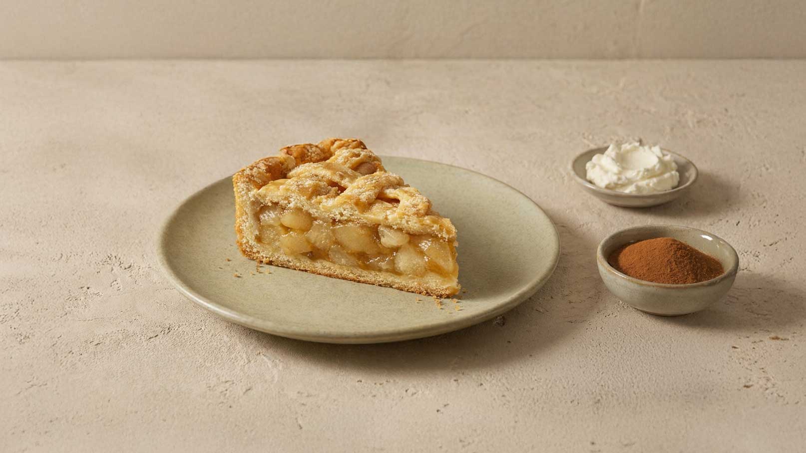 Ein Stück Apfelkuchen auf einem flachen Teller, umgeben von kleine Schalen mit Sahne und Zimt. Der Kuchen ist goldbraun und hat eine rustikale, geflochtene Kruste. Der Hintergrund ist neutral und schlicht.
