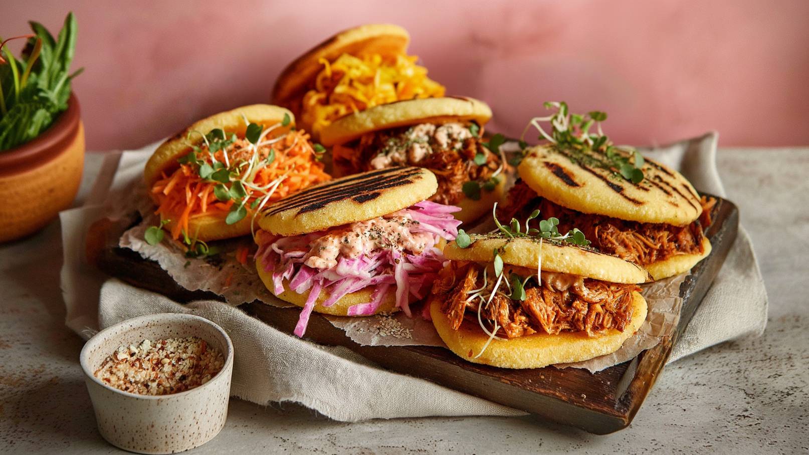 Sechs gefüllte Arepas auf einem Holzbrett, jede mit unterschiedlichen Zutaten wie geraspeltem Gemüse und Fleisch, garniert mit frischen Kräutern. Daneben ein kleiner Schüssel mit Gewürzen. Der Hintergrund ist sanft rosa, und die Präsentation ist ansprechend und einladend.