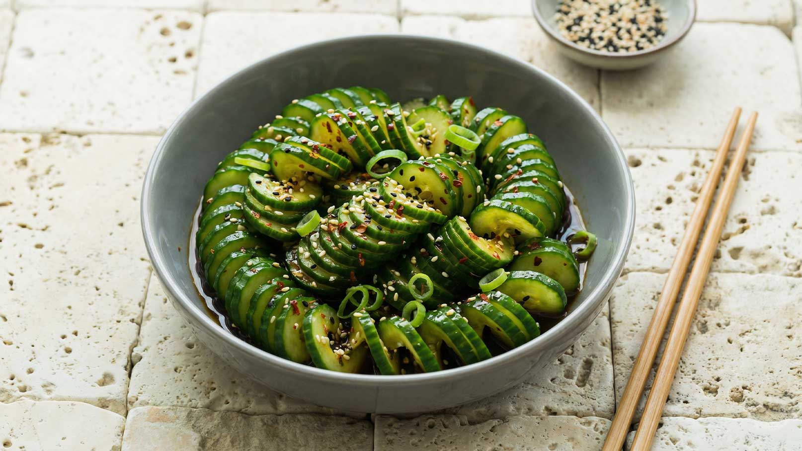 Ein grauer Schüssel enthält in Scheiben geschnittene Gurken, die ansprechend angerichtet sind. Die Gurken sind mit Frühlingszwiebeln und Sesamsamen dekoriert. Im Hintergrund liegen Essstäbchen.