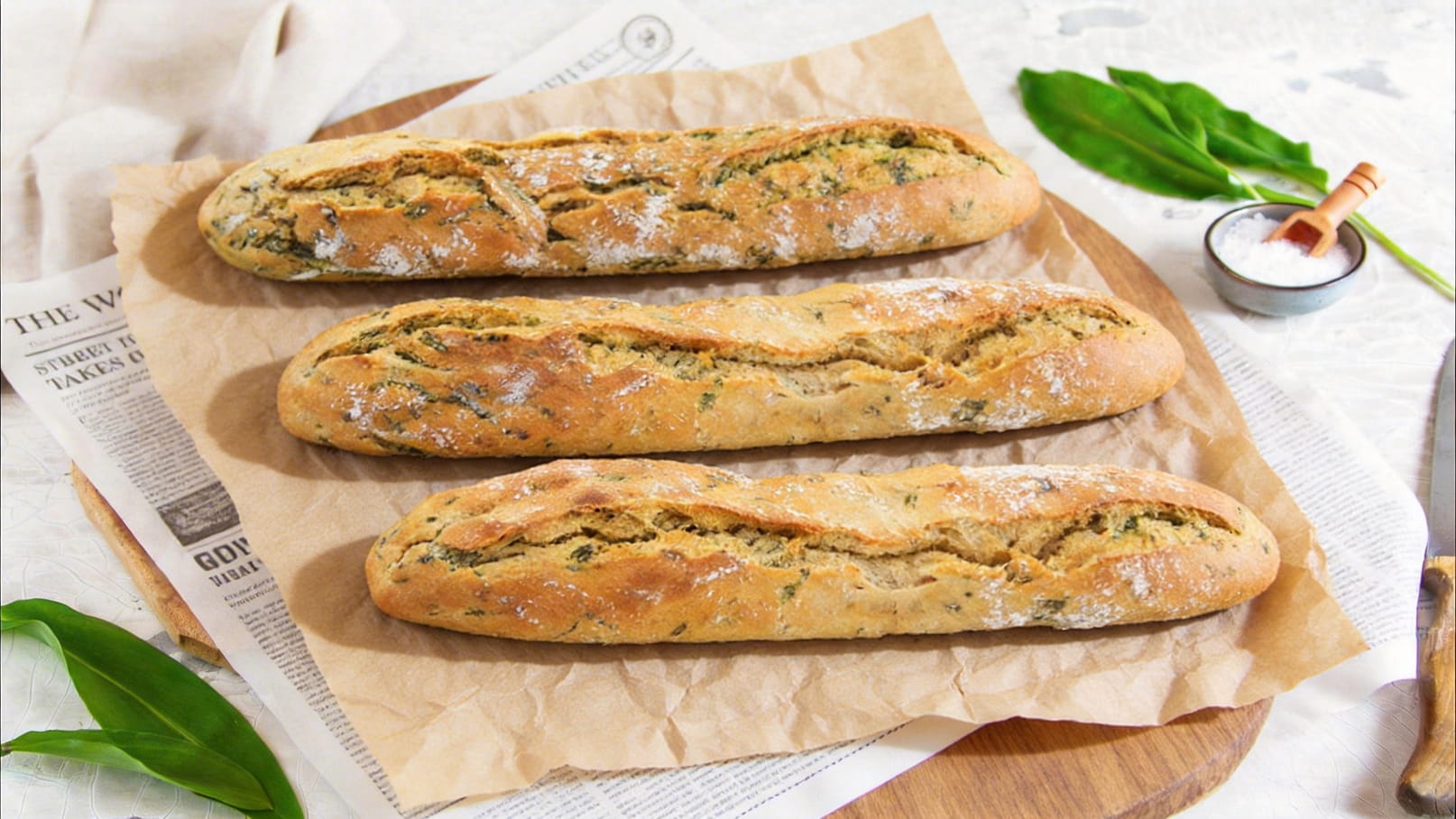 Drei frische, knusprige Baguettes liegen auf braunem Backpapier. Die Brote sind mit Kräutern verfeinert und haben eine goldbraune Kruste. Im Hintergrund sind dekorative Elemente wie Blätter und eine kleine Schüssel mit Salz zu sehen.