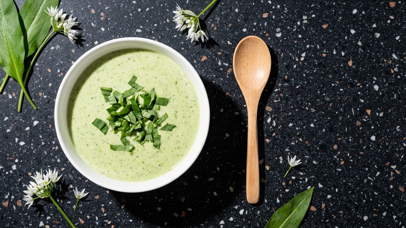 Eine Schüssel mit grüner Suppe ist im Mittelpunkt, garniert mit gehacktem Blattwerk. Neben der Schüssel liegt ein Holzlöffel. Um die Schüssel herum sind einige Pflanzenblätter und Blumen verteilt. Der Hintergrund ist dunkel mit einer körnigen Oberfläche.