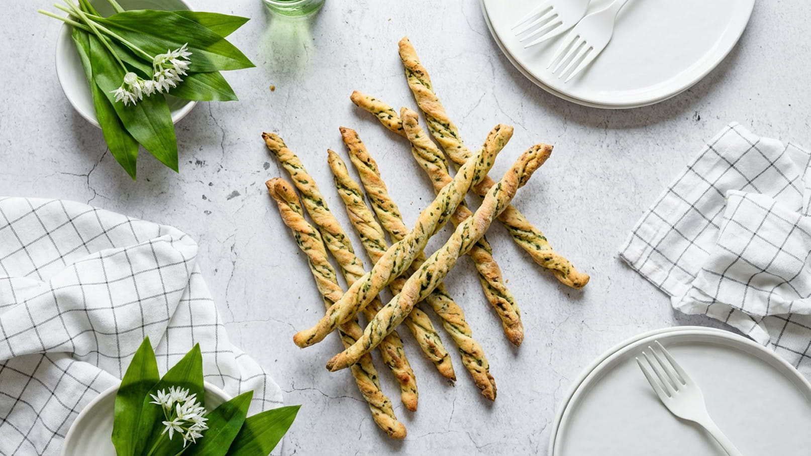 Auf dem Bild sind mehrere knusprige, spiralförmige Brote auf einem Tisch angeordnet. Um sie herum liegen zwei weiße Tellern und frische Blätter, die eine einladende, frühlingshafte Atmosphäre schaffen.