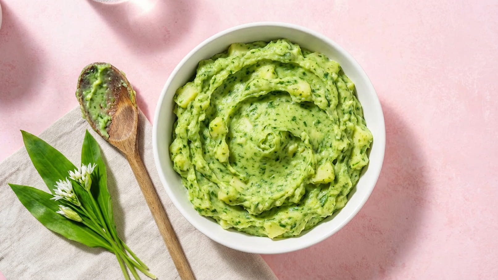 Eine weiße Schüssel enthält einen cremigen grünen Aufstrich, eventuell aus pürierten Kräutern und Gemüse. Daneben liegt ein Holzlöffel, der teilweise mit dem Aufstrich bedeckt ist. Im Vordergrund sind einige frische Blätter zu sehen. Der Hintergrund ist rosa.