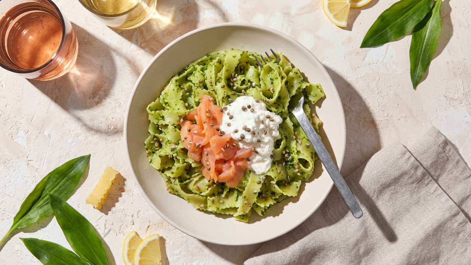 Ein Teller mit grüner Pasta, garniert mit Räucherlachs und einer Portion Ricotta. Ein Besteck ist daneben platziert. Im Hintergrund sind Gläser mit Getränken und Zitronenscheiben sichtbar. Frische Blätter und ein Stück Käse ergänzen das Gesamtbild.