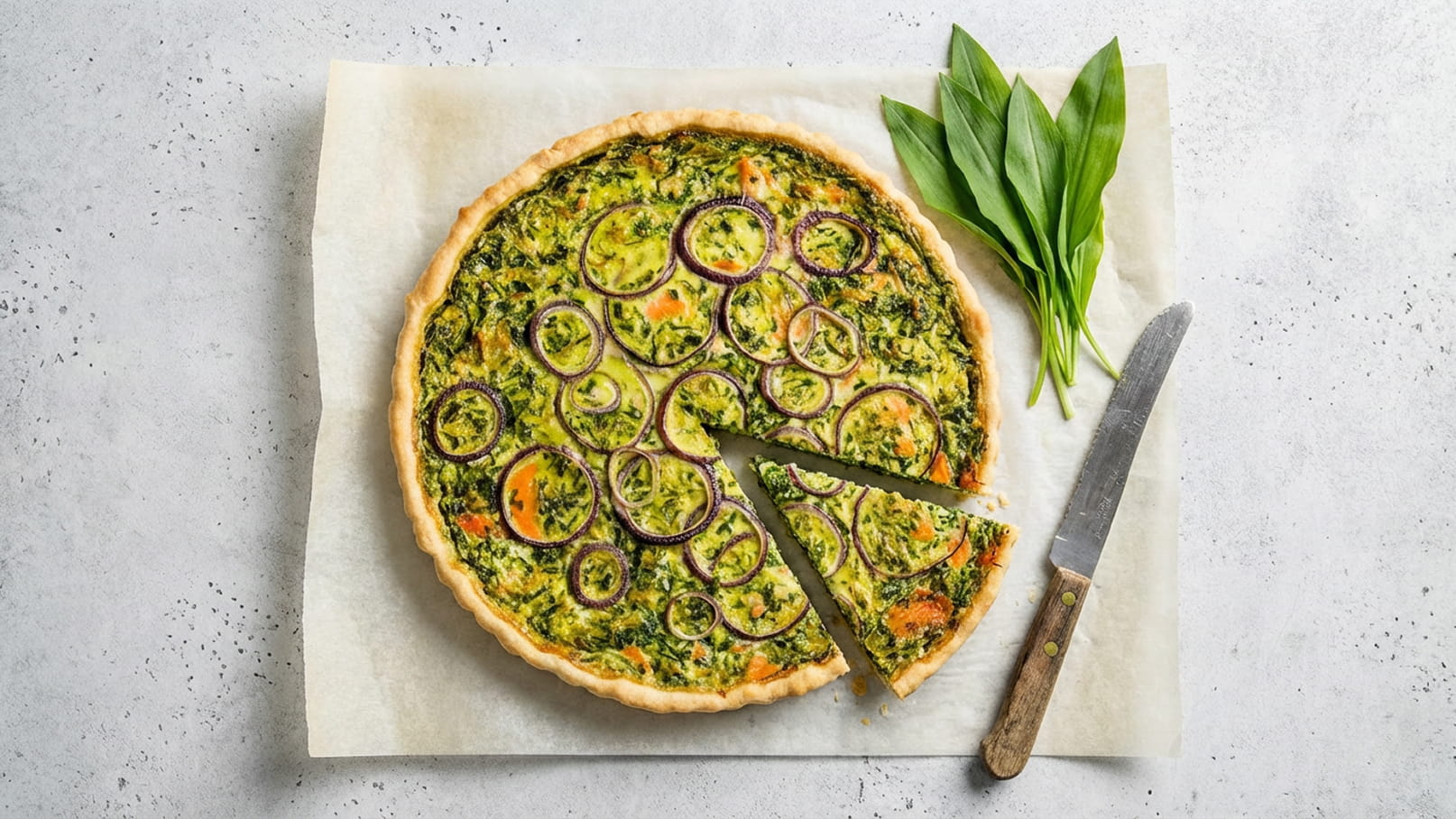 Eine Quiche mit einer bunten Füllung aus Blattgemüse, Zucchini und lila Zwiebeln. Ein Stück ist bereits herausgeschnitten. Neben der Quiche liegen frische Bärlauchblätter und ein Messer. Der Hintergrund ist hell und neutral.