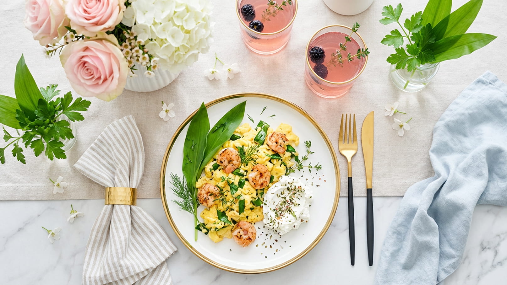 Ein Teller mit Rührei, Kräutern und Garnelen steht auf einem eleganten Tisch. Daneben befinden sich drei Gläser mit pinken Getränken, dekoriert mit Beeren, und eine Blumenvase mit Rosen und weißen Blumen. Ein dekoratives Serviettenbündel und Besteck ergänzen die Tischdekoration.