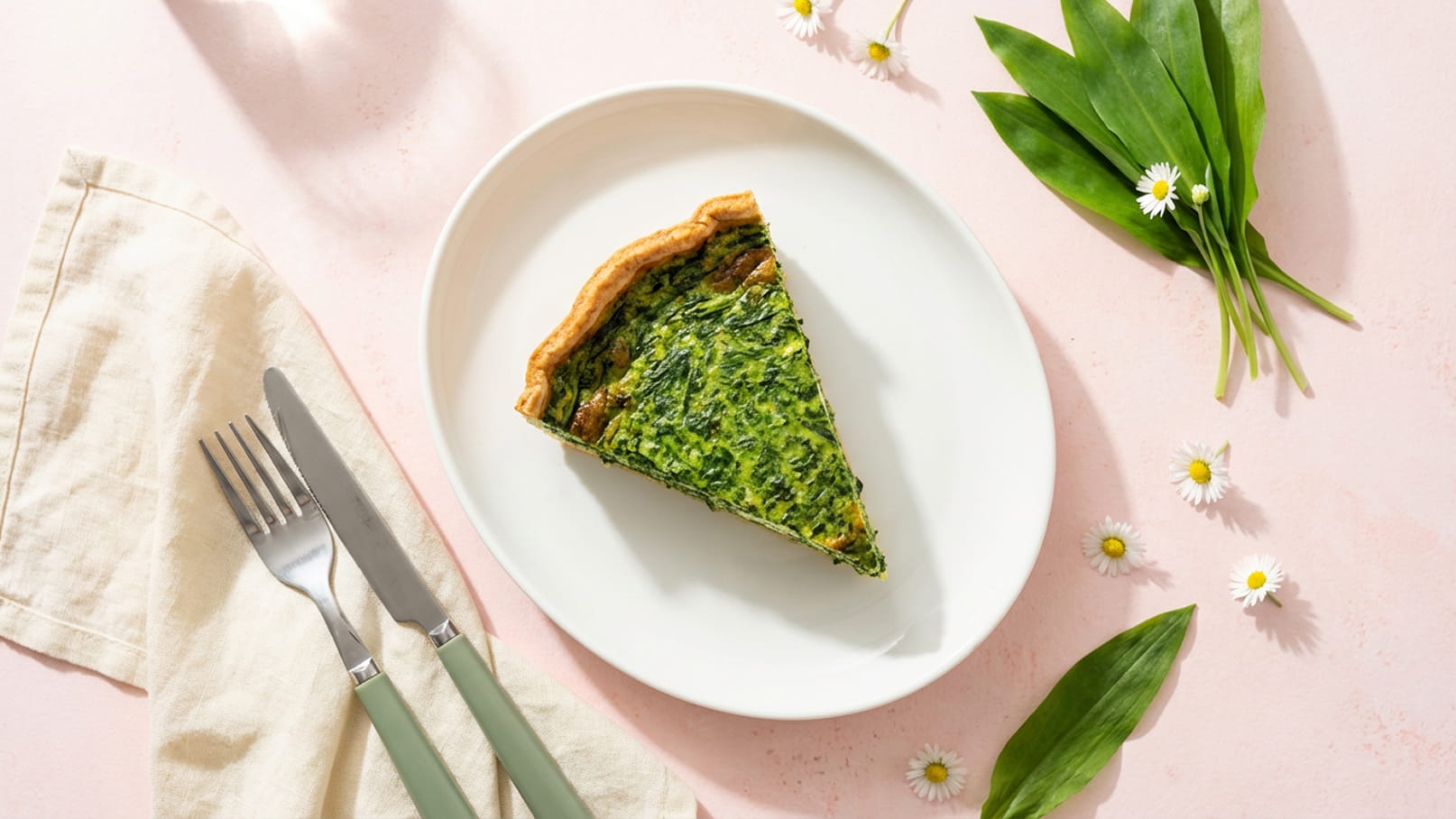Auf dem Bild ist ein Stück grüne Quiche auf einem weißen Teller zu sehen. Der Teig ist goldbraun, und die Füllung besteht aus Blattgemüse. Der Teller liegt auf einer rosa Unterlage, umgeben von frischen Kräutern und kleinen Blumen.