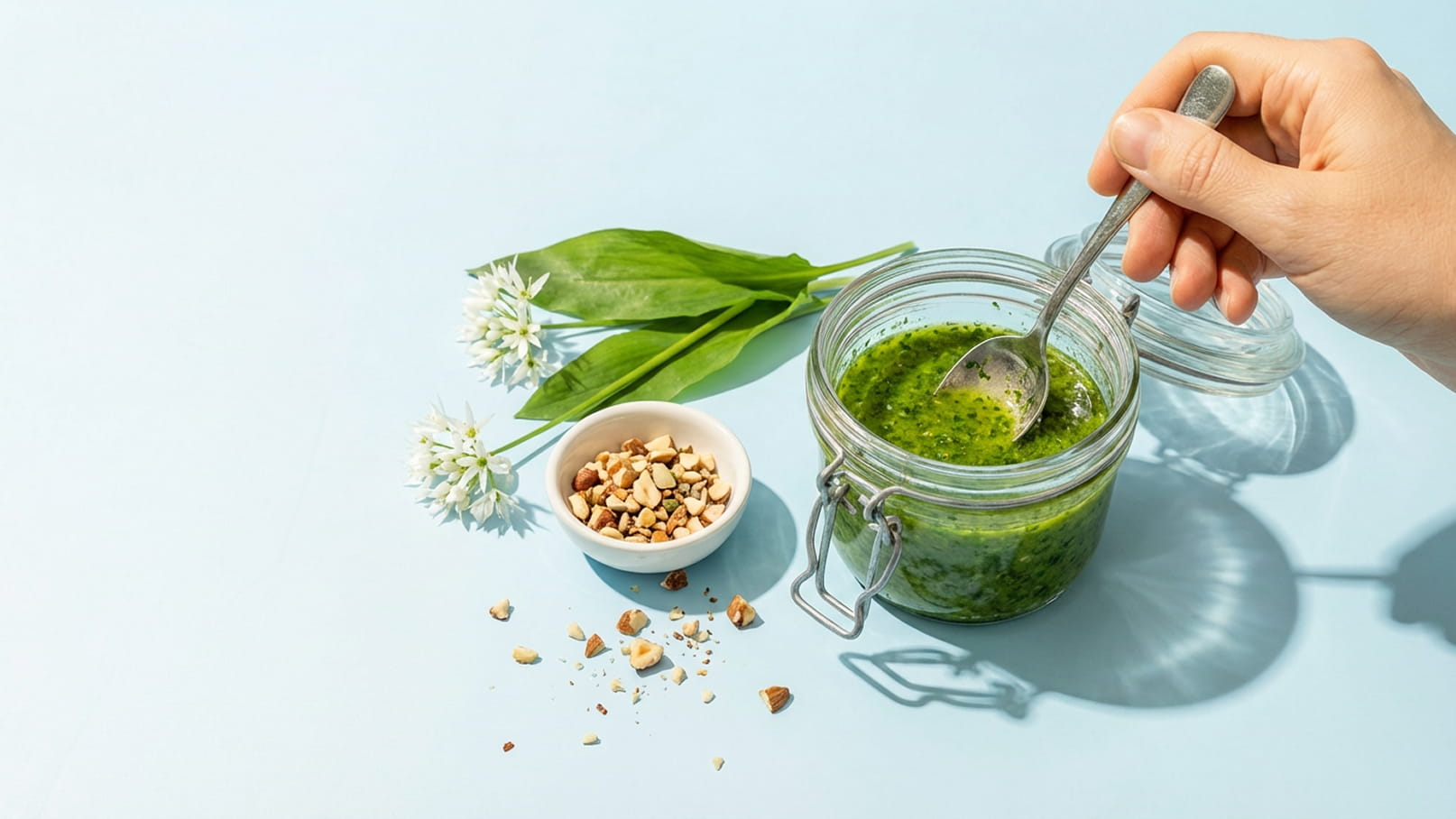 Eine Hand hält einen Löffel und rührt in einem Glas mit grünem Pesto. Neben dem Glas befindet sich eine Schale mit gehackten Nüssen und einige frische Bärlauchblätter. Der Hintergrund ist hellblau.