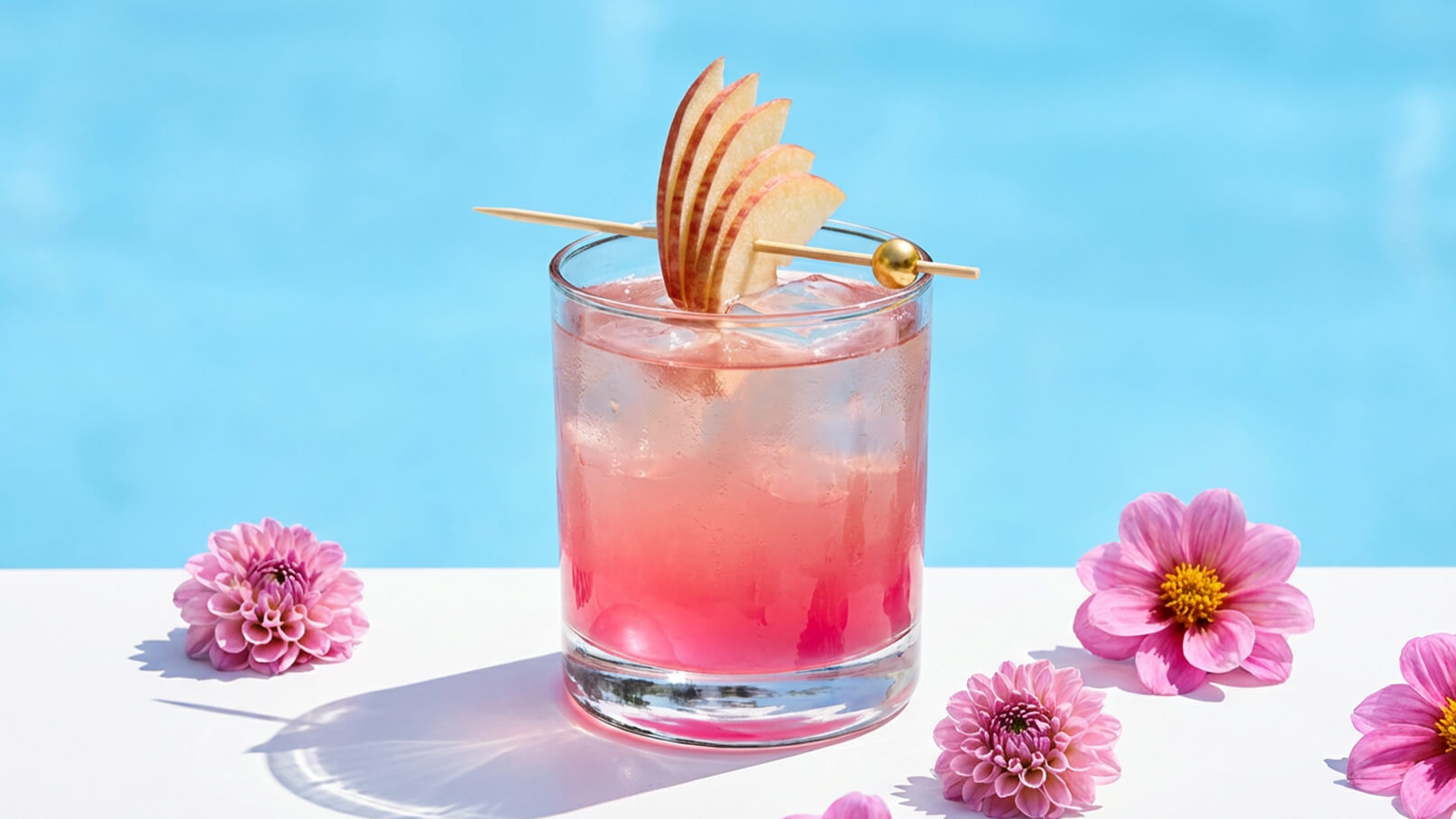 Ein Cocktail in einem Glas mit Eiswürfeln und einem fruchtigen, rosafarbenen Getränk. Oben garnierte Apfelscheiben und ein eleganter Zahnstocher. Um das Glas herum liegen ein paar pinkfarbene Blumen. Der Hintergrund zeigt einen blauen Pool.