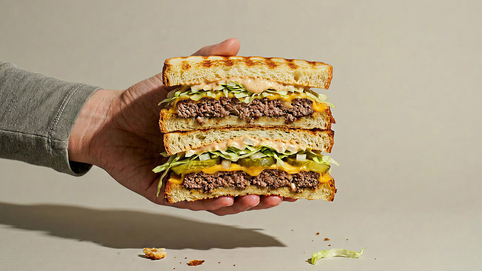 Eine Hand hält einen großen, mehrschichtigen Hamburger. Die Schichten bestehen aus gegrilltem Fleisch, geschmolzenem Käse, frischem Salat, Gurkenscheiben und einer Sauce, alles zwischen zwei gerösteten Brötchen. Im Hintergrund ist ein neutraler Farbton zu sehen.