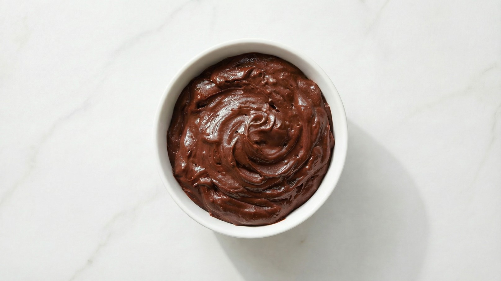 Eine weiße Schüssel enthält eine glatte, cremige Schokoladenmasse mit einer glänzenden Oberfläche. Die Masse scheint dick und reichhaltig zu sein, ideal für Desserts oder als Belag. Der Hintergrund ist hell und unauffällig.