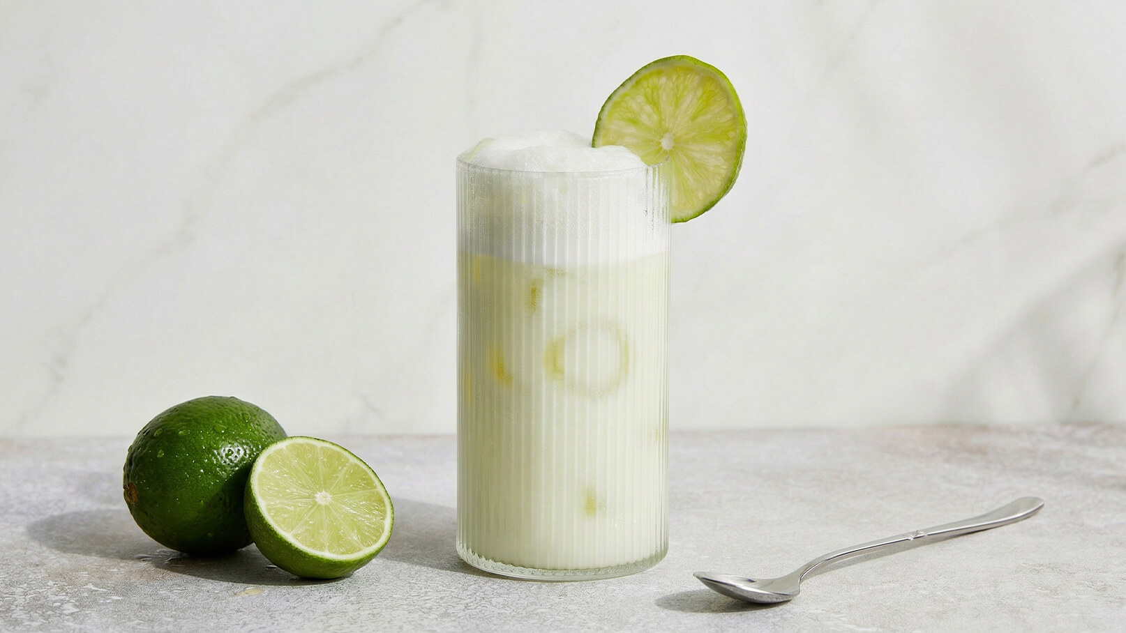 Ein hoher, transparenter Becher mit einem erfrischenden Getränk, verziert mit einer Limettenscheibe. Neben dem Becher liegen zwei frischeLimetten. Der Hintergrund ist hell und neutral, was den Fokus auf das Getränk legt.
