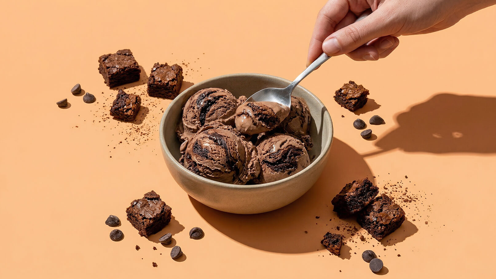 Eine Hand hält einen Löffel und schöpft Schokoladeneis aus einer runden Schüssel. Um die Schüssel herum liegen Stücke von Brownies und Schokoladendrops. Der Hintergrund ist in einem warmen Orangeton gehalten.