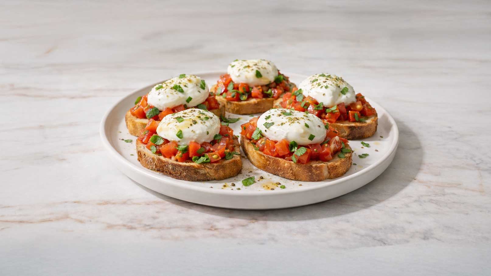 Ein runder Teller mit vier Bruschetta. Jedes Stück Brot ist mit einer Mischung aus gewürfelten Tomaten, Kräutern und einer Portion pochiertem Ei belegt. Die Bruschetta sind mit frischen Kräutern garniert. Der Teller steht auf einer hellen Oberfläche.