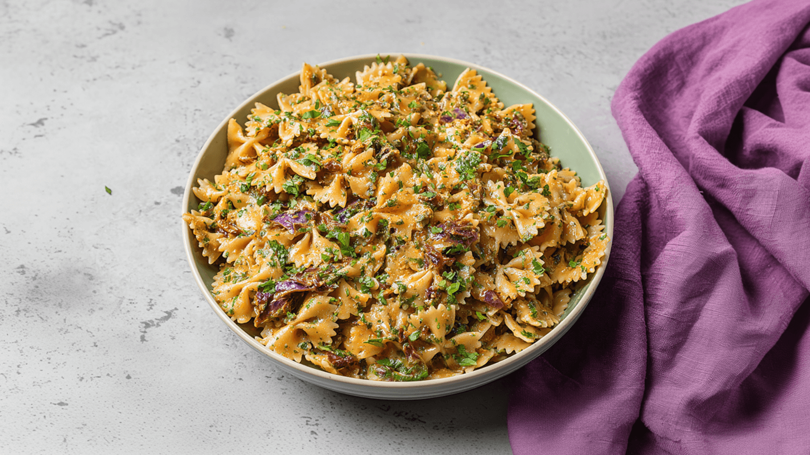 Eine Schüssel mit farfalle Pasta ist gefüllt mit einer cremigen Sauce und reichlich frischem, gehacktem Koriander. Die Pasta ist gleichmäßig mit Sauce umhüllt und einige Stücke roter Paprika sind sichtbar. Im Hintergrund liegt ein lila Tuch.