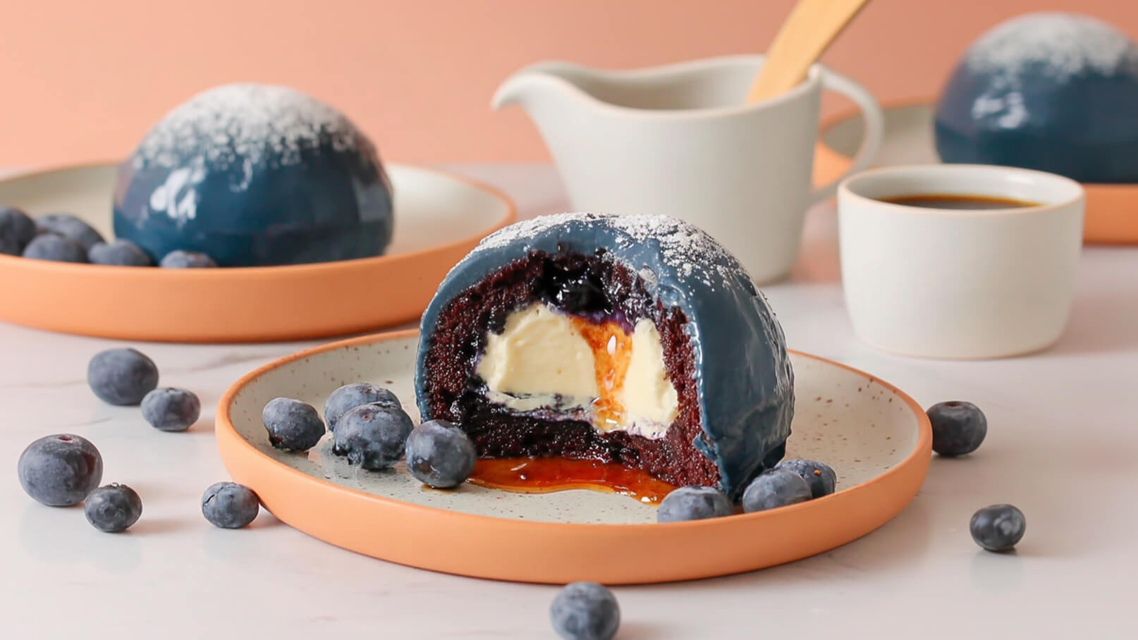 Auf dem Bild ist ein aufgeschnittenes Dessert zu sehen, das aus einem runden, dunklen Kuchen mit einer glänzenden blauen Glasur besteht. Im Inneren befinden sich eine cremige Füllung und eine fruchtige Komponente. Um den Teller sind frische Heidelbeeren verteilt.