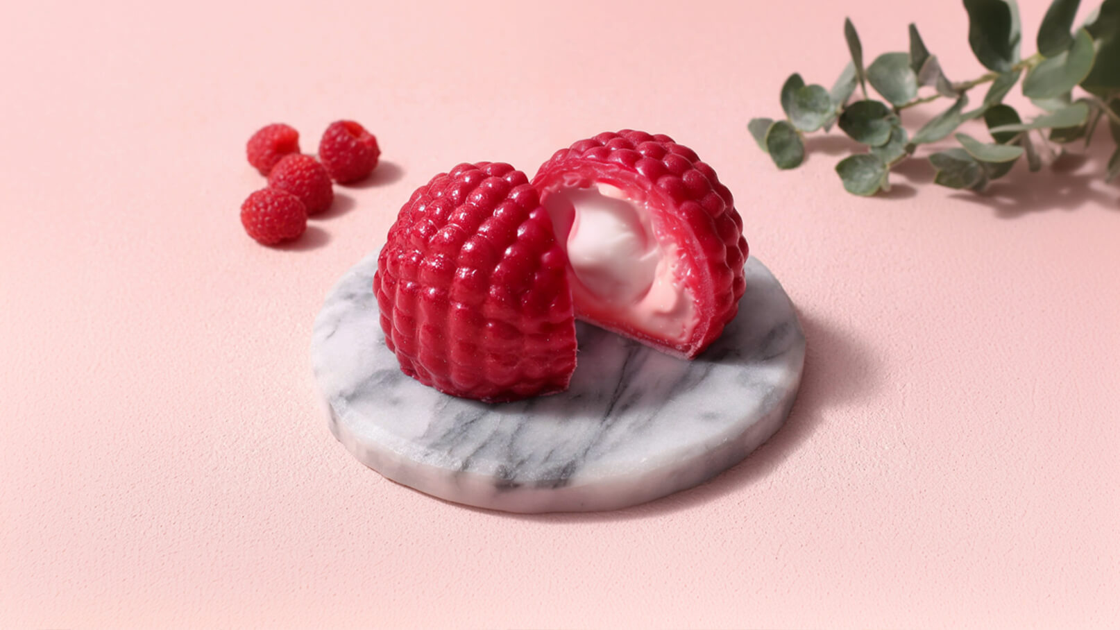 Auf einem marmornen Teller liegt eine halbe, rote Himbeerkonfektion, die eine cremige Füllung zeigt. Neben der Konfektion befinden sich frische Himbeeren. Der Hintergrund ist in sanften Pastellfarben gehalten.