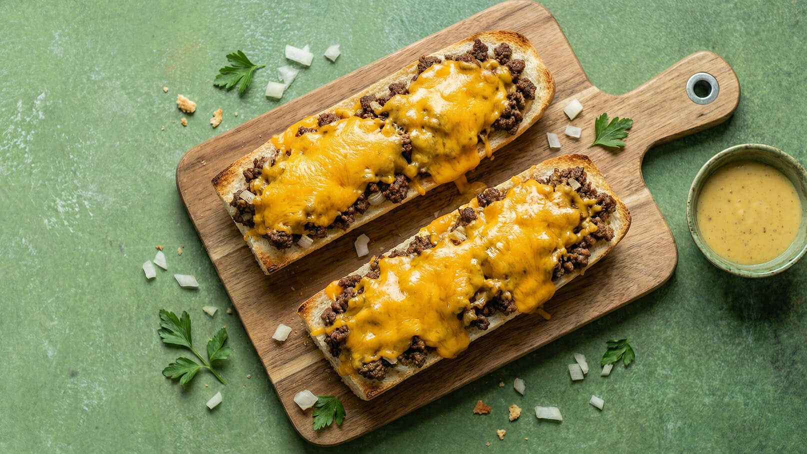Zwei belegte Baguettes auf einem Holzbrett, gefüllt mit einer Mischung aus Hackfleisch und reichlich geschmolzenem Käse. Dazu eine Schüssel mit einer gelben Sauce. Die Umgebung ist grün und mit frischen Kräutern dekoriert.