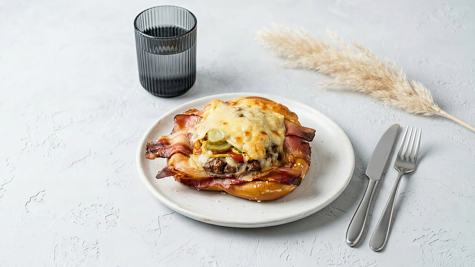 Ein Teller mit einem herzhaften Brötchen belegt mit Schinken, geschmolzenem Käse und Gurken. Daneben steht ein Glas mit einem dunklen Getränk und Besteck liegt bereit.