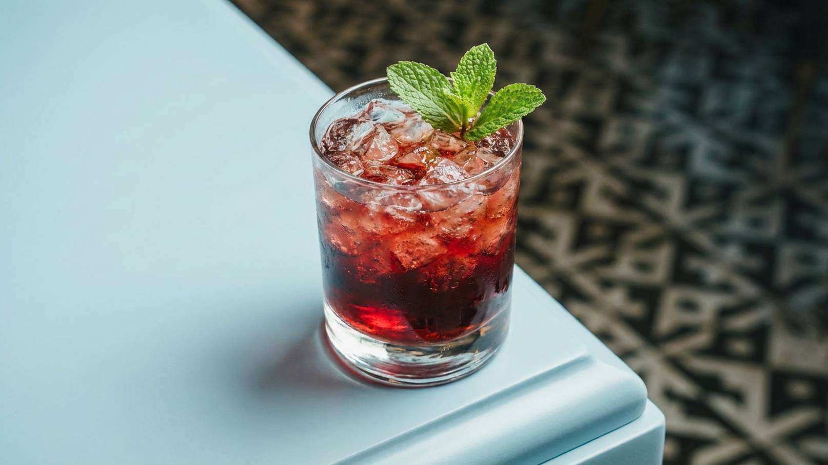 Ein Glas mit einem roten Getränk, gefüllt mit Eiswürfeln, steht auf einem Tisch. Oben wird das Getränk von frischen Minzblättern garniniert.
