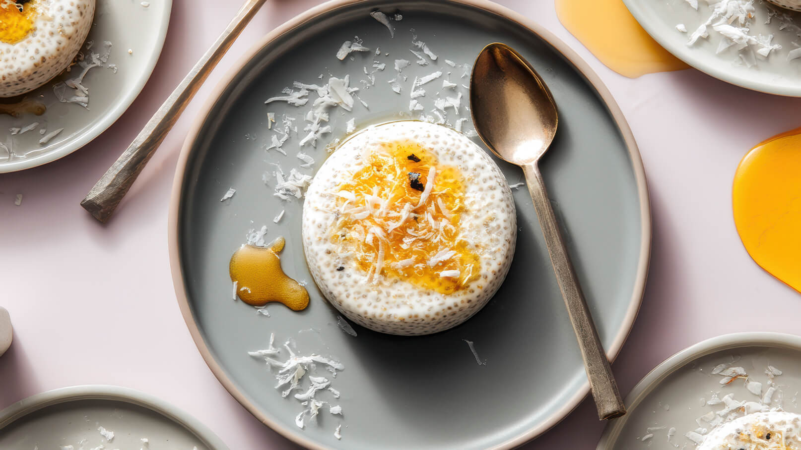 Ein rundes Dessert in einem grauen Teller, garniert mit Kokosraspeln und einem orangenen Topping. Neben dem Teller liegt ein Löffel aus Metall. Einige weitere Teller mit ähnlichem Inhalt sind im Hintergrund zu sehen.
