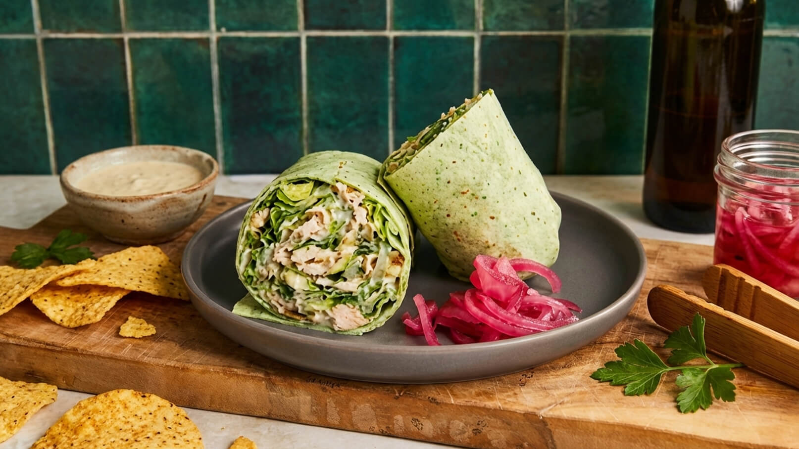 Auf dem Bild sind zwei aufgerollte Wraps auf einem grauen Teller zu sehen, gefüllt mit Gemüse und Hähnchen. Daneben liegt eine Portion eingelegter roter Zwiebeln. Im Hintergrund gibt es Tortilla-Chips und eine kleine Schüssel mit Dipsauce.