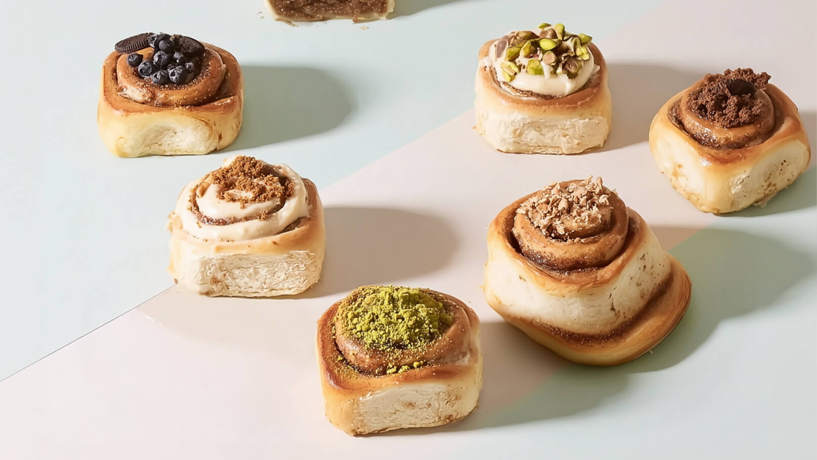 Eine Auswahl von Zimtschnecken mit verschiedenen Toppings auf einem hellen Hintergrund. Die Schnecken sind in verschiedenen Geschmacksrichtungen wie Schokolade, Pistazie und Kekskrümel dekoriert, was eine vielfältige und ansprechende Süßigkeit bietet.