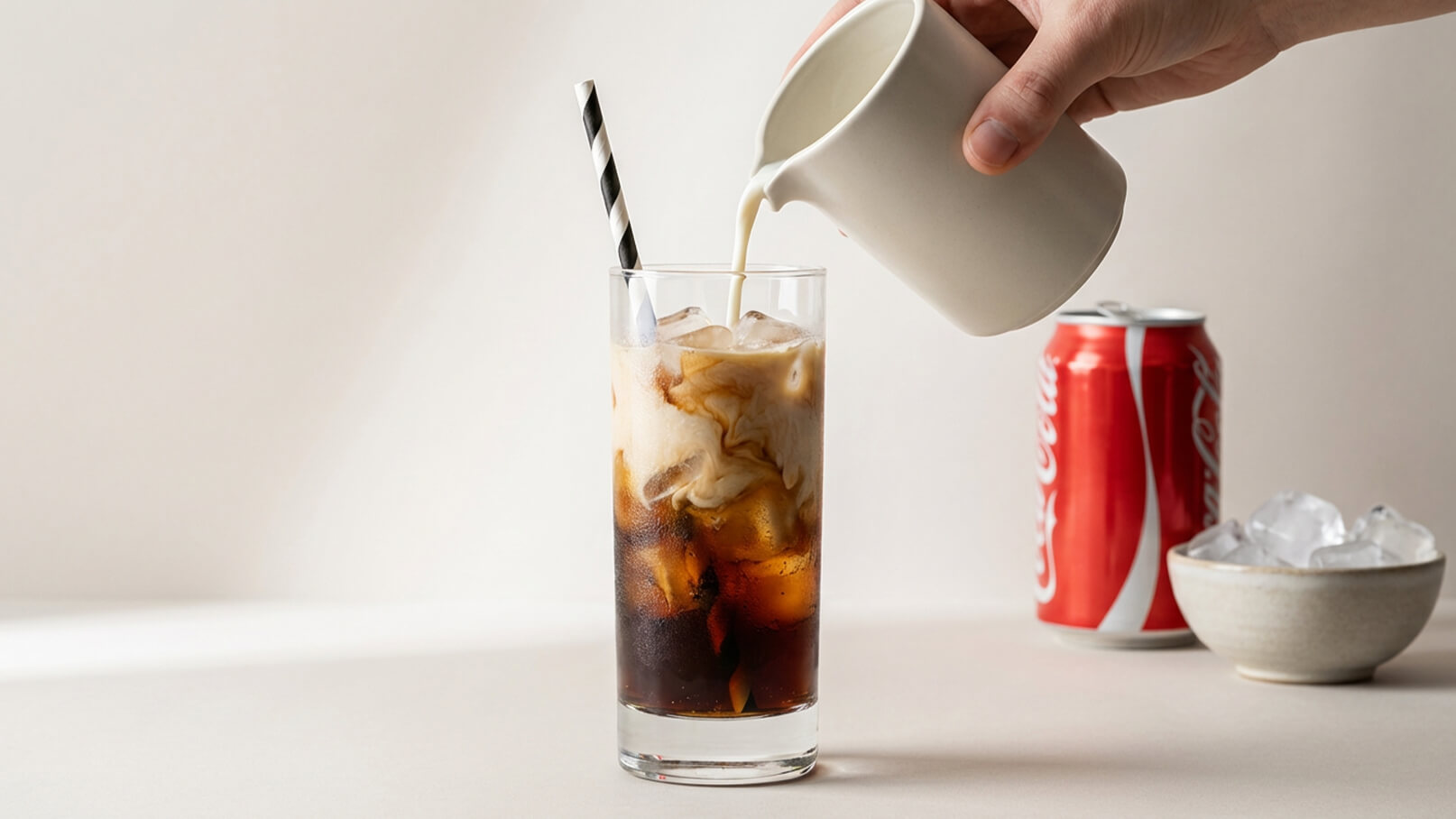 Ein Glas mit Eis und einer Mischung aus Kaffee und Milch wird gerade von einer Hand mit einer Kanne Milch übergossen. Im Hintergrund sind eine Schale mit Eiswürfeln und zwei Dosen Cola sichtbar.