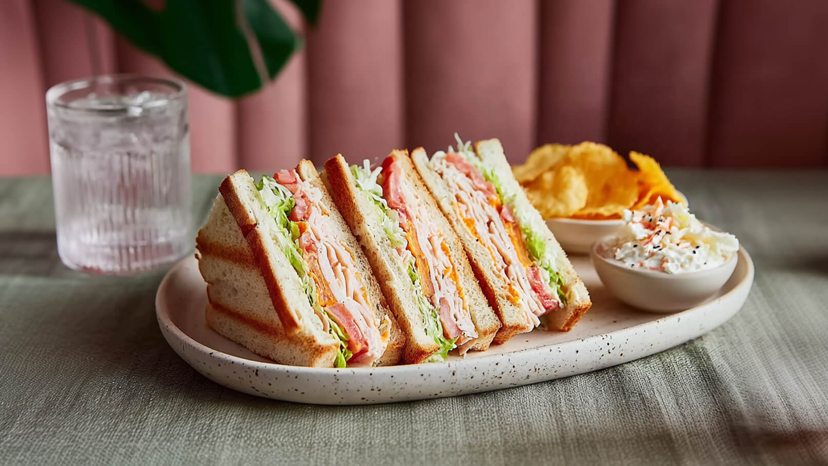 Ein Teller mit mehreren Schichten Sandwiches, gefüllt mit Pute, Käse, Tomaten und Salat. Neben den Sandwiches befinden sich Kartoffelchips und eine kleine Schüssel mit Krautsalat. Im Hintergrund steht ein Glas Wasser.