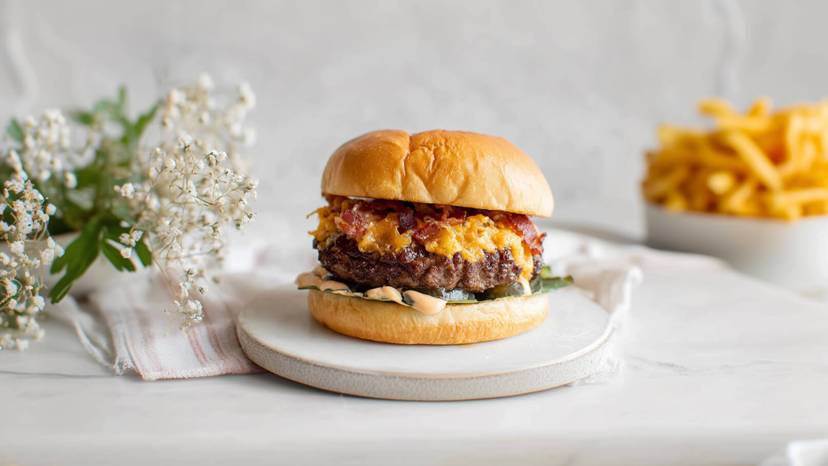 Ein saftiger Hamburger auf einem weißen Teller, belegt mit geschmolzenem Käse, Speck und einer Salatgurkenscheibe. Er ist umgeben von einem weichen Brötchen. Im Hintergrund sind frische Blumen und eine Schüssel mit Pommes frites sichtbar.