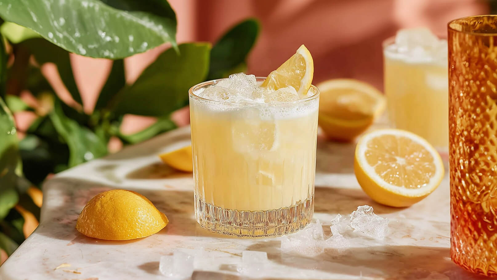 Ein Glas erfrischenden, gelben Limonade mit Eiswürfeln und einer Zitronenscheibe dekoriert steht auf einem marmorierten Tisch. Im Hintergrund liegen einige Zitronenhälften und grüne Blätter sind sichtbar.