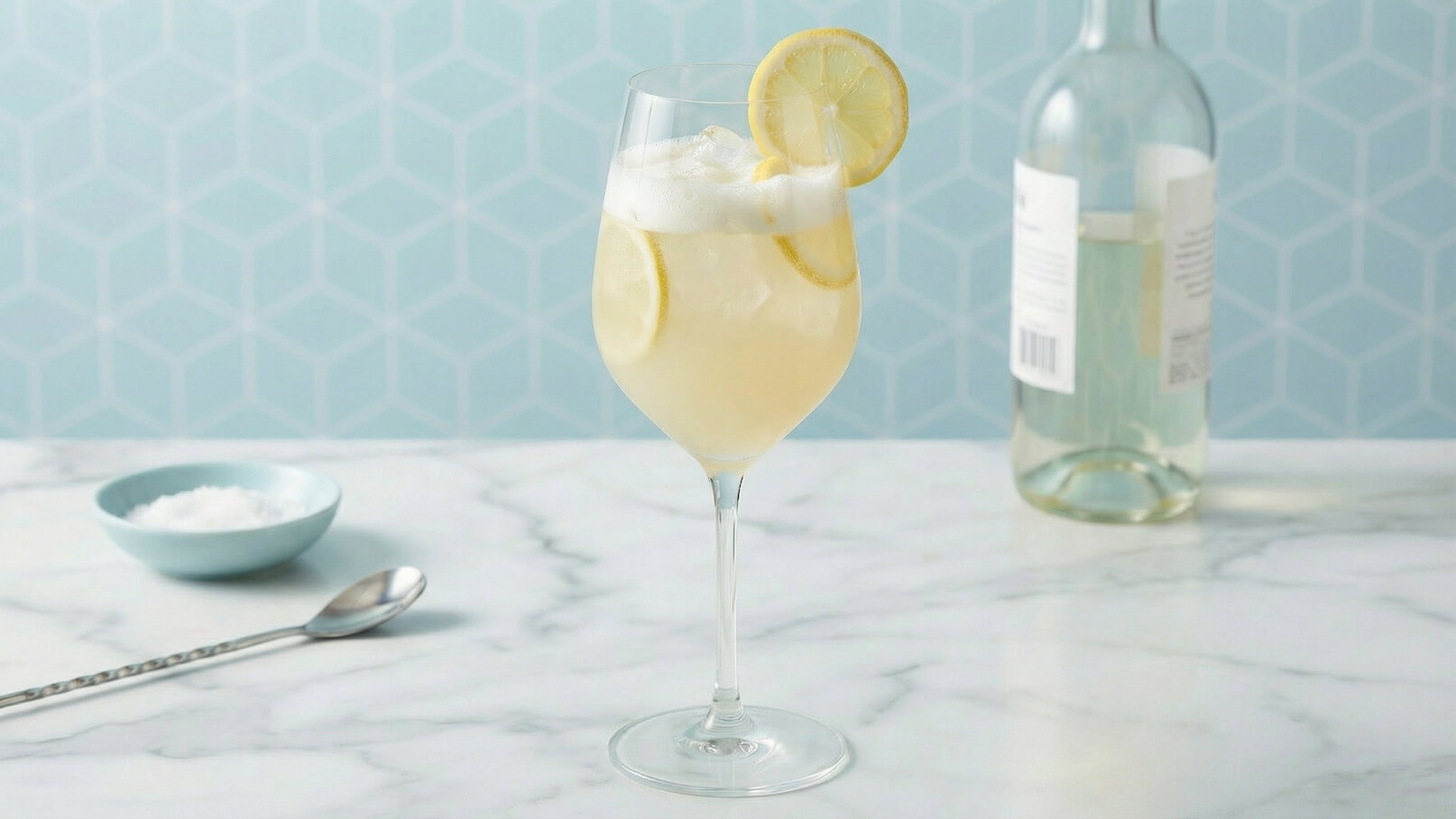 Ein Glas mit einem sprudelnden Getränk, das mit Zitronenscheiben garniert ist. Im Hintergrund steht eine Flasche. Auf dem Tisch befindet sich eine kleine Schüssel mit Salz und ein Löffel. Der Hintergrund ist in einem hellen, geometrischen Muster gehalten.