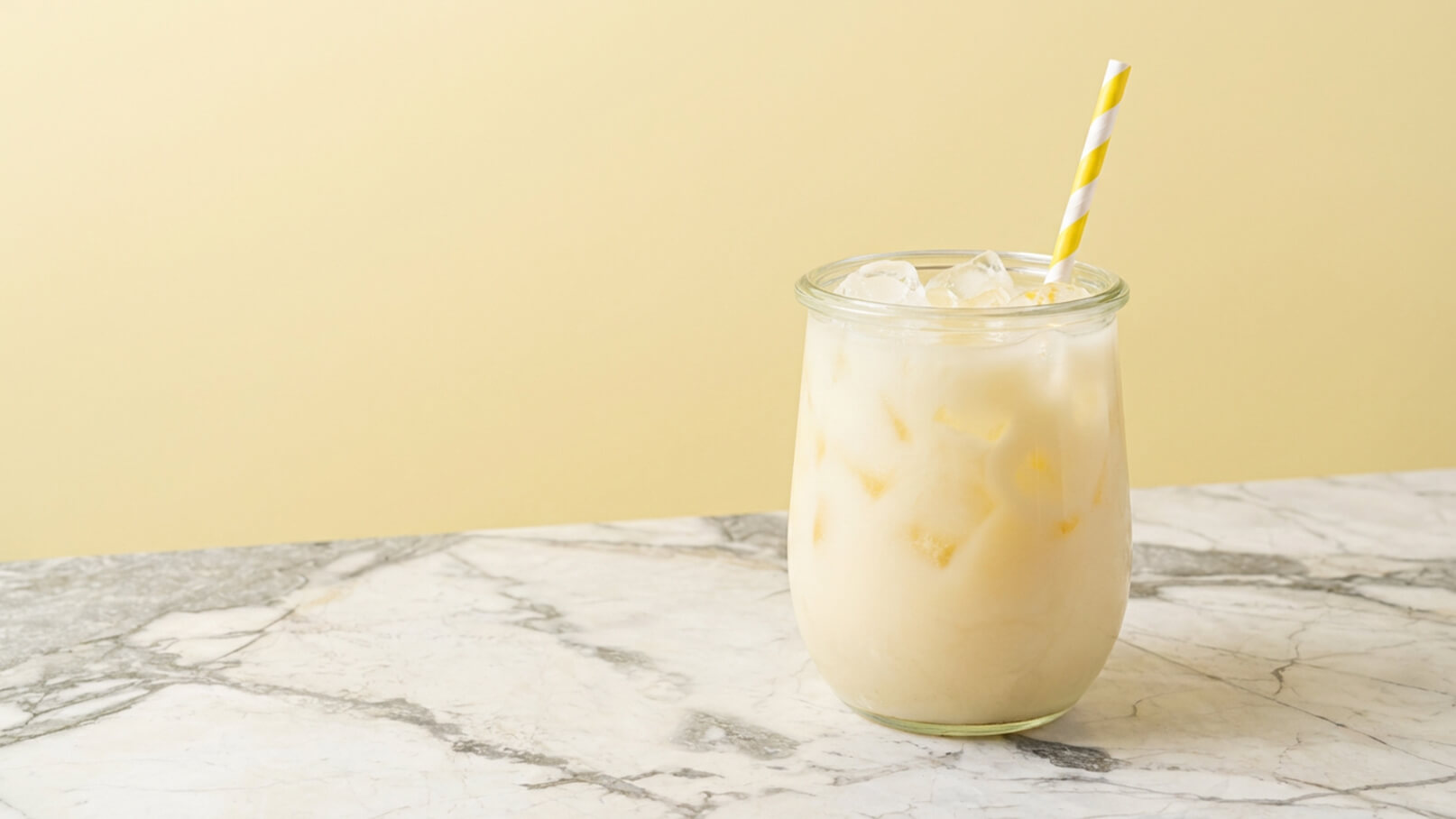Ein Glas mit einem erfrischenden, weißen Getränk ist auf einem Marmor-Tisch platziert. Das Glas enthält Eiswürfel und hat einen gelb-weiß gestreiften Strohhalm. Der Hintergrund ist in sanften Gelbtönen gehalten.