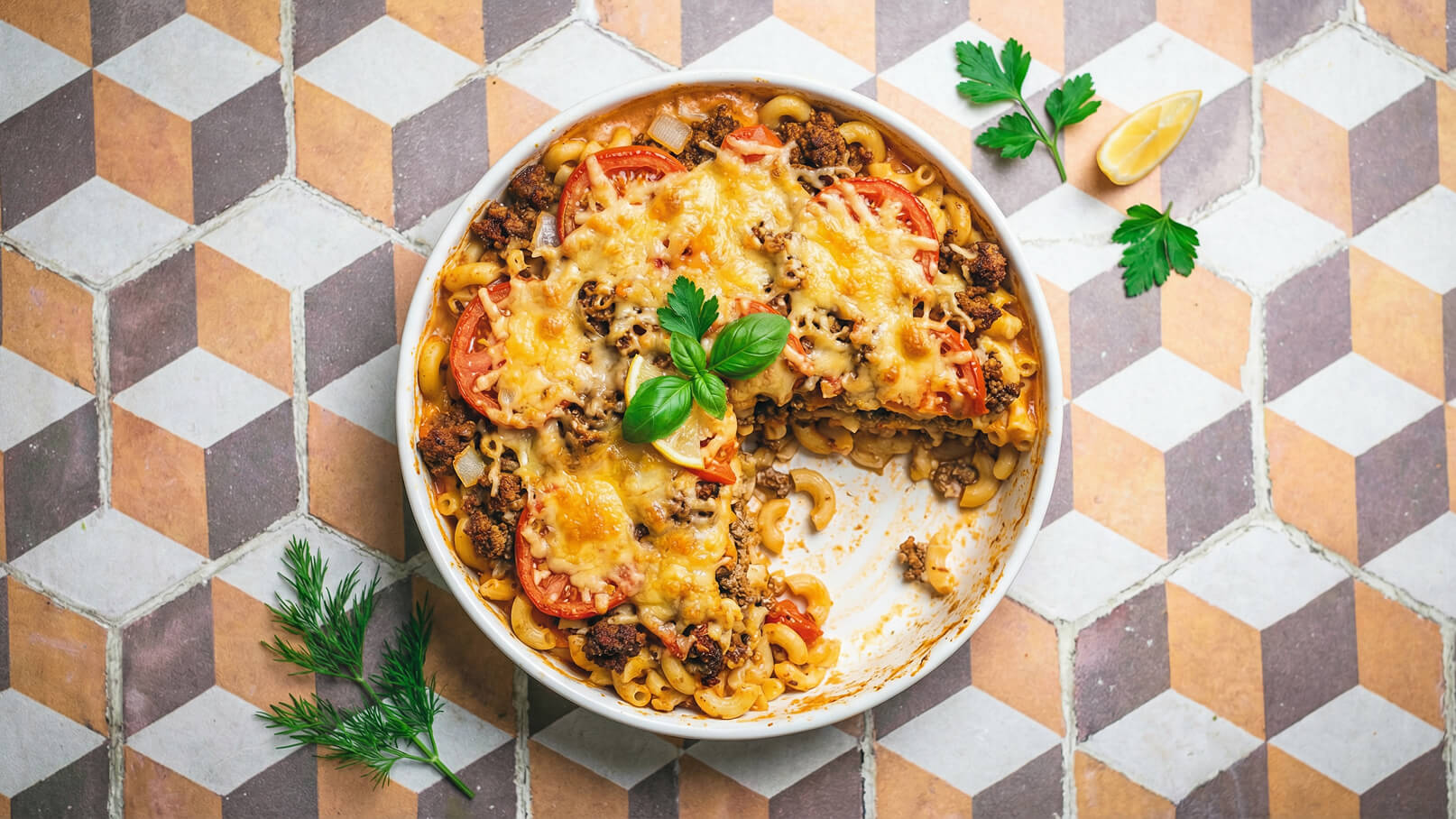 Ein Auflauf aus Makkaroni, Hackfleisch und Tomaten ist dekorativ in einer runden Auflaufform angerichtet. Der Auflauf ist goldbraun überbacken und mit frischen Kräutern, darunter ein Basilikumblatt, garniert. Teile des Auflaufs wurden bereits entnommen.