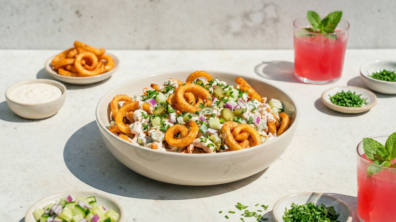 Eine große Schüssel mit Salat, garniert mit knusprigen Spiralen, roten Zwiebeln und Gurkenstücken. Um die Schüssel herum stehen kleine Schalen mit Dips und frischen Kräutern sowie zwei Gläser mit einer pinken Getränk und Minzblättern.