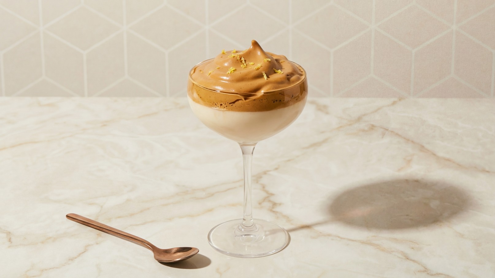 Ein elegantes Dessert in einem hohen Glas. Der untere Teil besteht aus einer cremigen Schicht, während die Oberfläche mit einem schaumigen, hellbraunen Topping verziert ist. Neben dem Glas liegt ein esslöffelförmiger Löffel.