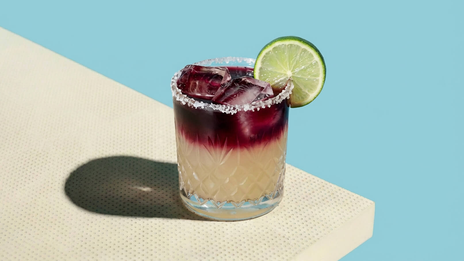 Ein Glas mit einem bunten Cocktail steht auf einem Tisch. Der Drink hat eine Schicht mit lila Farbe oben und eine hellere Schicht darunter. Der Rand des Glases ist mit Salz verziert, und eine Limettenscheibe hängt am Rand. Eiskü cubes sind sichtbar im Glas.