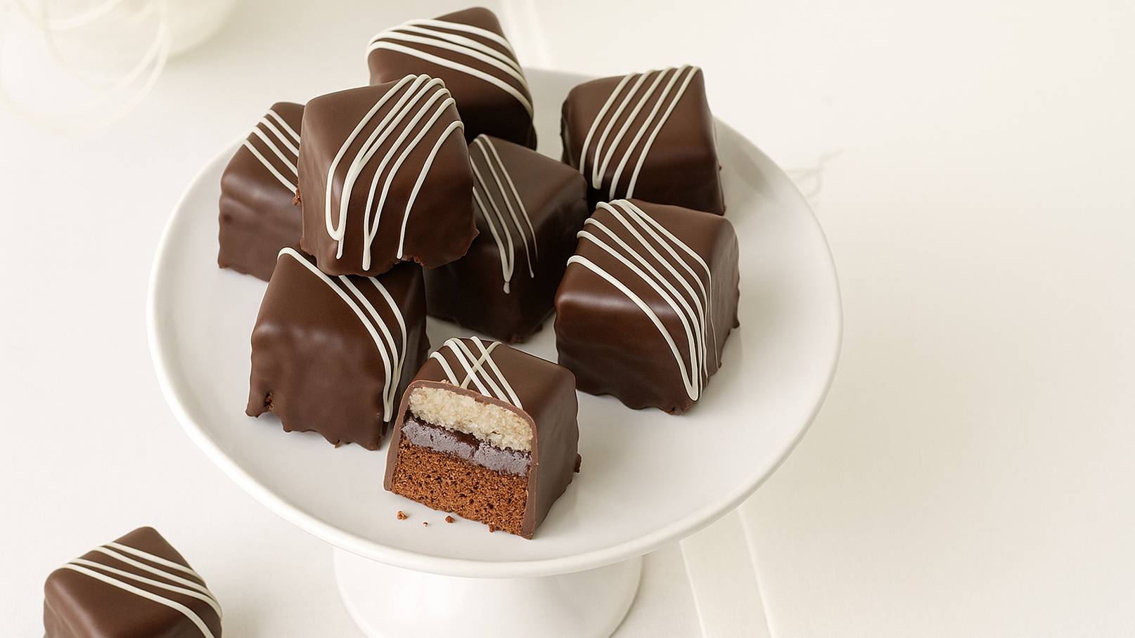 Ein Dessertteller mit mehreren klassisch quadratischen Pralinen, überzogen mit dunkler Schokolade und dekoriert mit weißen Schokoladengittern. Eine Praline ist aufgeschnitten, sodass die Schichten aus Biskuit, Schokoladencreme und einer nussigen Füllung sichtbar sind.