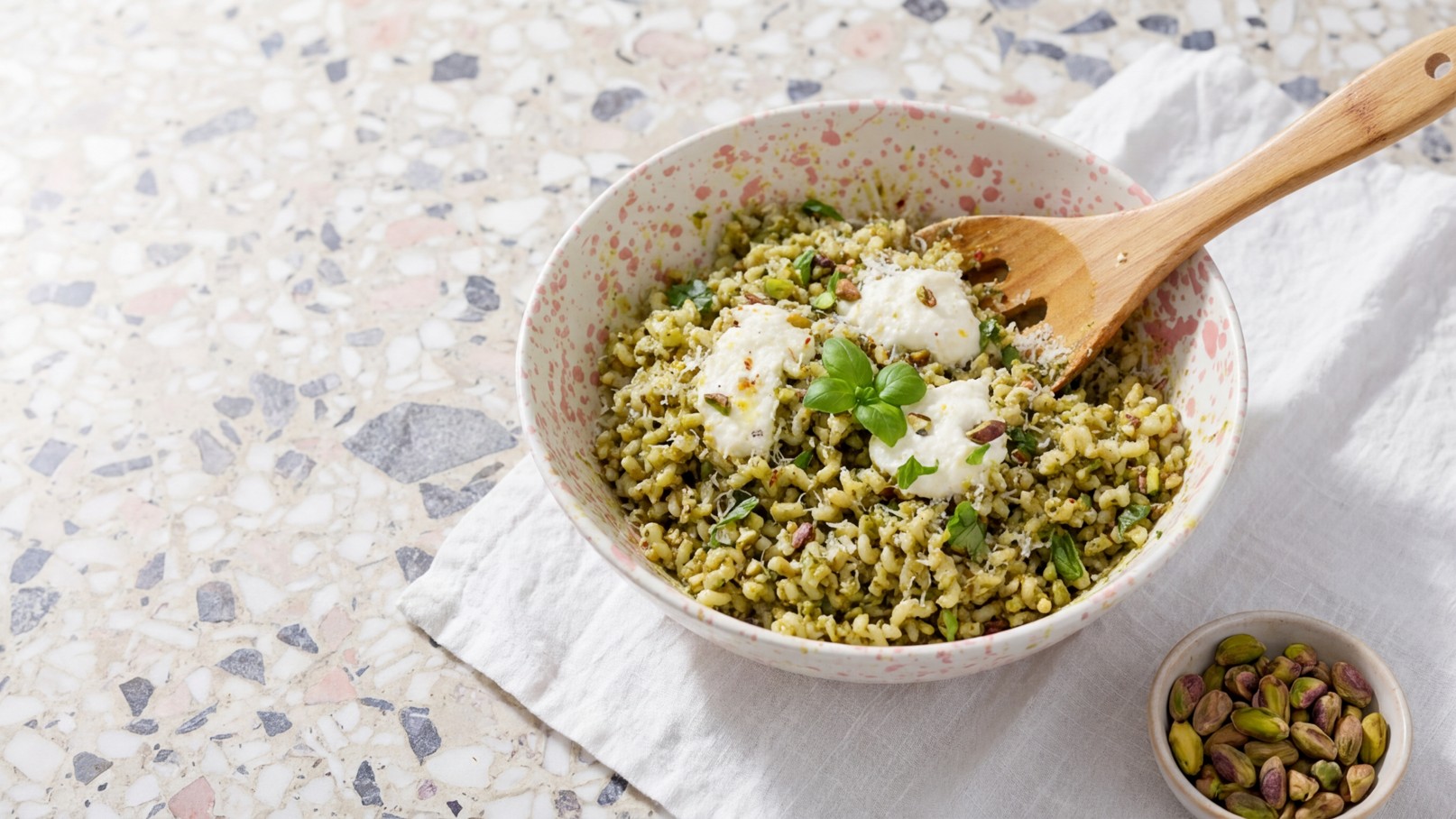 Eine Schüssel mit grünem Salat aus Körnern, garniert mit Joghurt und frischen Kräutern, steht auf einem hellen Tisch. Neben der Schüssel befindet sich eine kleine Schale mit Pistazien. Ein Holzlöffel liegt in der Schüssel.