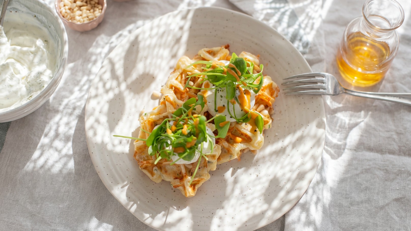 Ein Teller mit zwei Portionen einer warmen Speise, garniert mit frischem Gemüse und Kräutern. Neben dem Teller steht eine Schale mit Joghurt und ein Glas mit dressing. Die Szene wird von sanftem Licht durch ein Gittermuster beleuchtet.